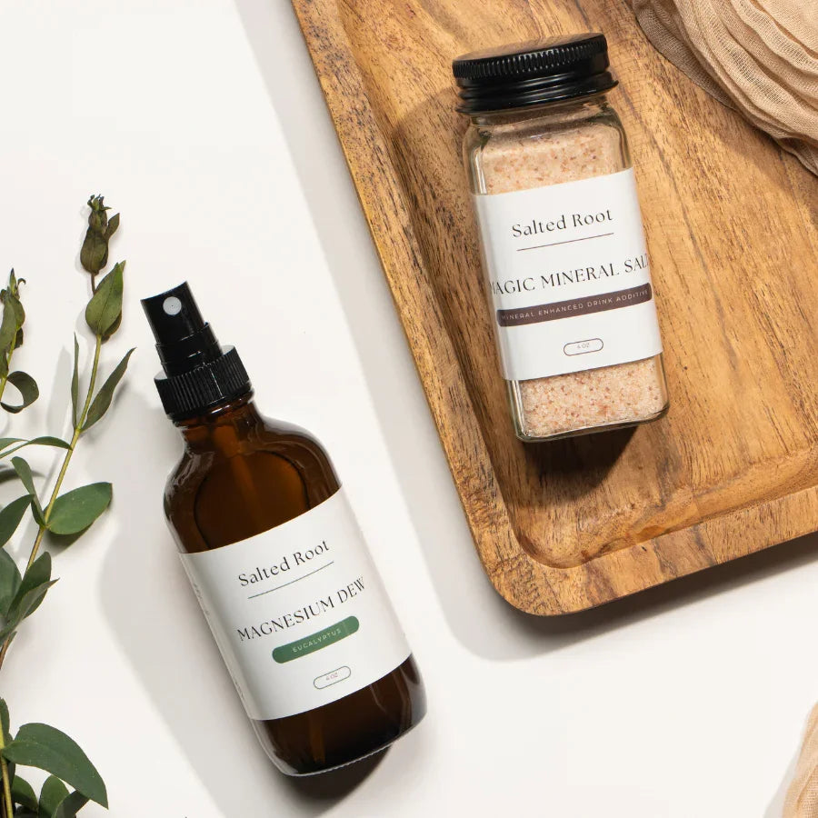 Flat lay of Salted Root products — Magic Mineral Salt in a glass jar placed on a wooden tray, and Magnesium Dew spray bottle lying beside it on a white surface with eucalyptus sprigs, evoking a clean, natural wellness aesthetic.